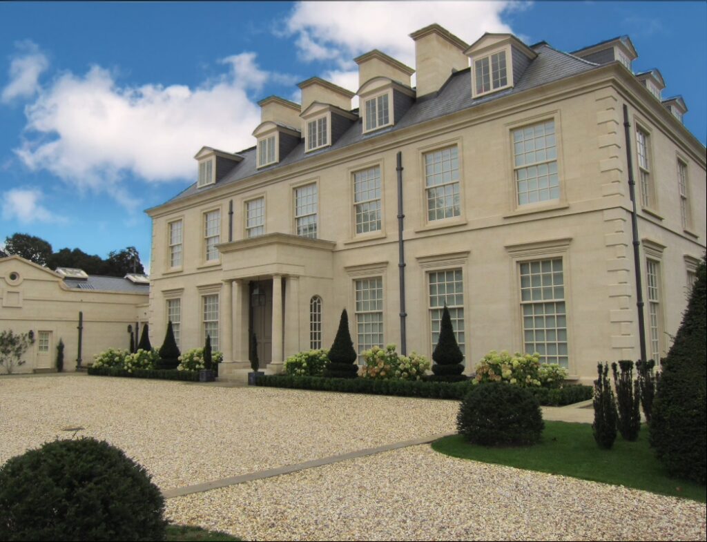 Large beige mansion with symmetrical windows, columned entrance, and a gravel driveway bordered by manicured shrubs and topiary trees.