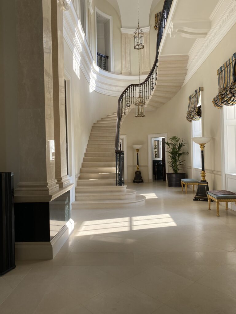 Grand foyer with a sweeping curved marble staircase, ornate black iron railing, and hanging chandeliers in a bright, classical interior.