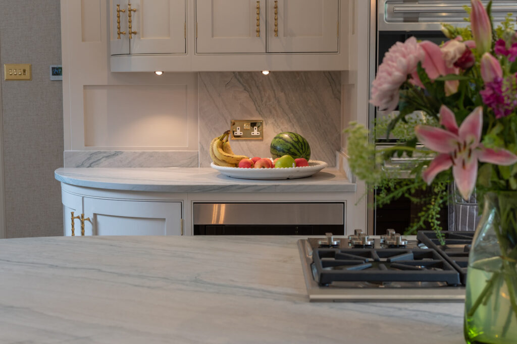 White marble kitchen island with a fruit bowl (bananas, apples, peaches and a watermelon) and a bright bouquet on the right.